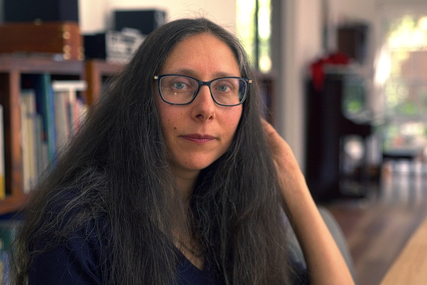 A woman with long dark hair and glasses sits at a table. She is looking at the camera with a neutral expression.