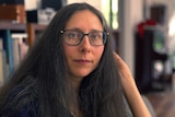 A woman with long dark hair and glasses sits at a table. She is looking at the camera with a neutral expression.