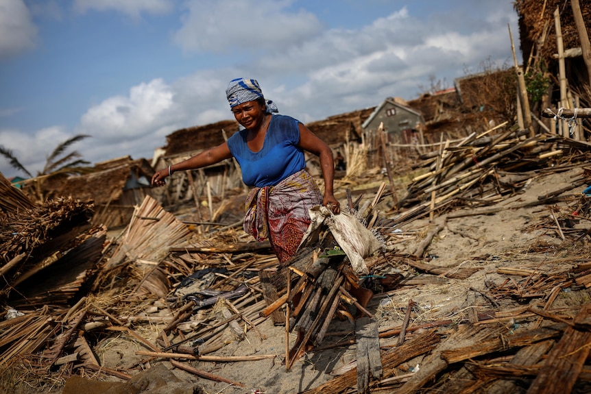 A woman carries wood, with debris in the background.