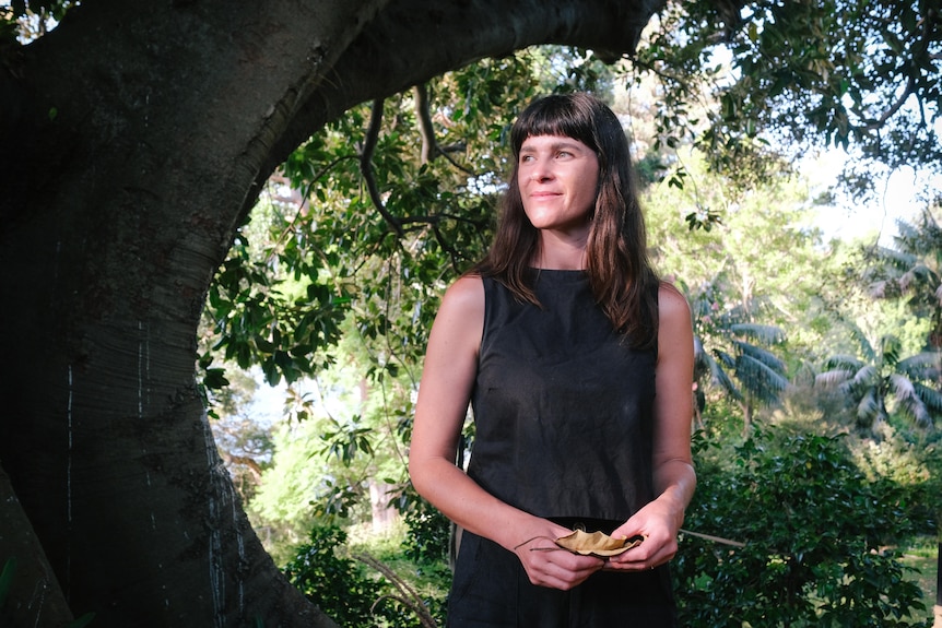 A brown haired 30-something womad stands under a large fig tree, smiling into the middle-distance
