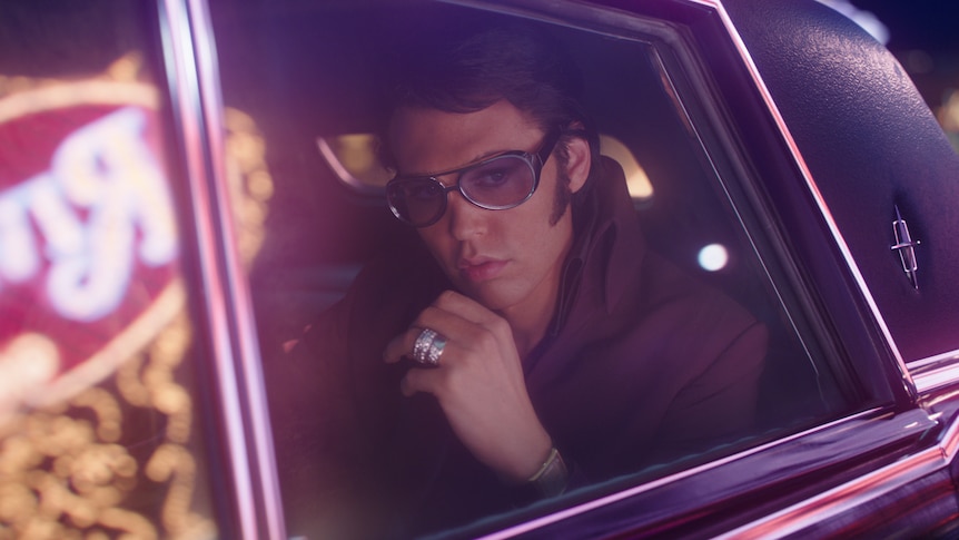 White man dressed as Elvis with 50s pompadour dark hair, sideburns and glitzy sunglasses sits looking out window of a Cadillac.