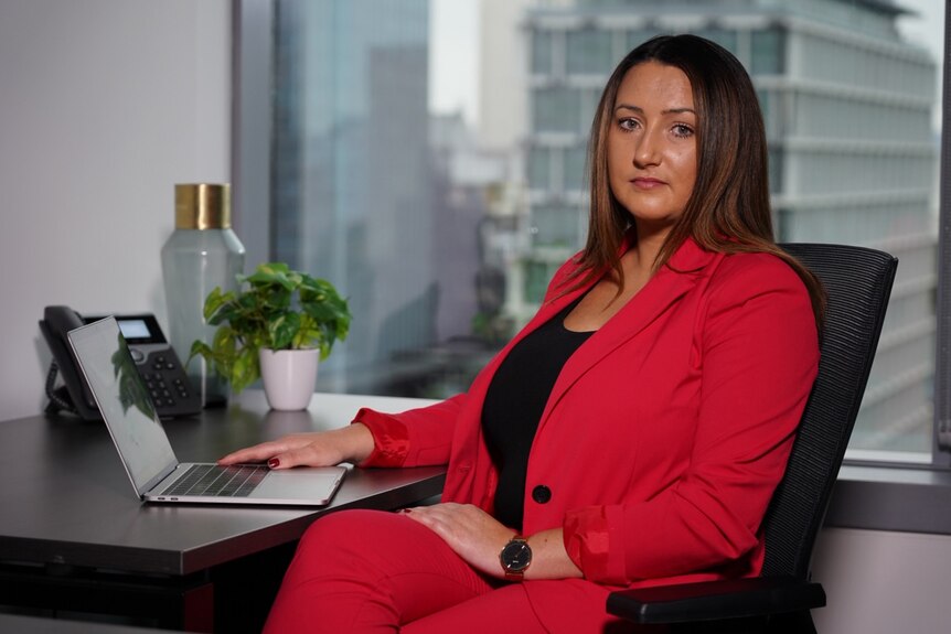 A woman in a red jacket and black top sits at a desk in an office in front of a laptop computer, posing for a photo.