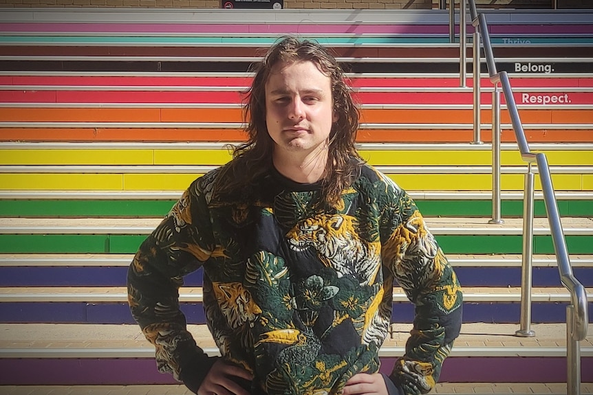 Mitch Evans stands with his hands on his hips, in front of a rainbow staircase.