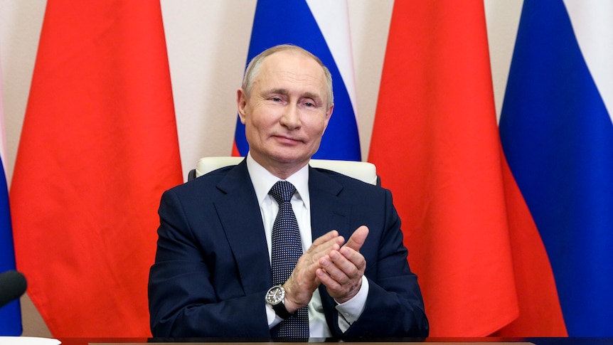 Vladimir Putin, sitting at a desk in front of Chinese and Russian flags, claps and smiles.