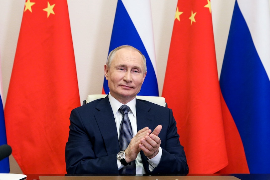 Vladimir Putin, sitting at a desk in front of Chinese and Russian flags, claps and smiles.