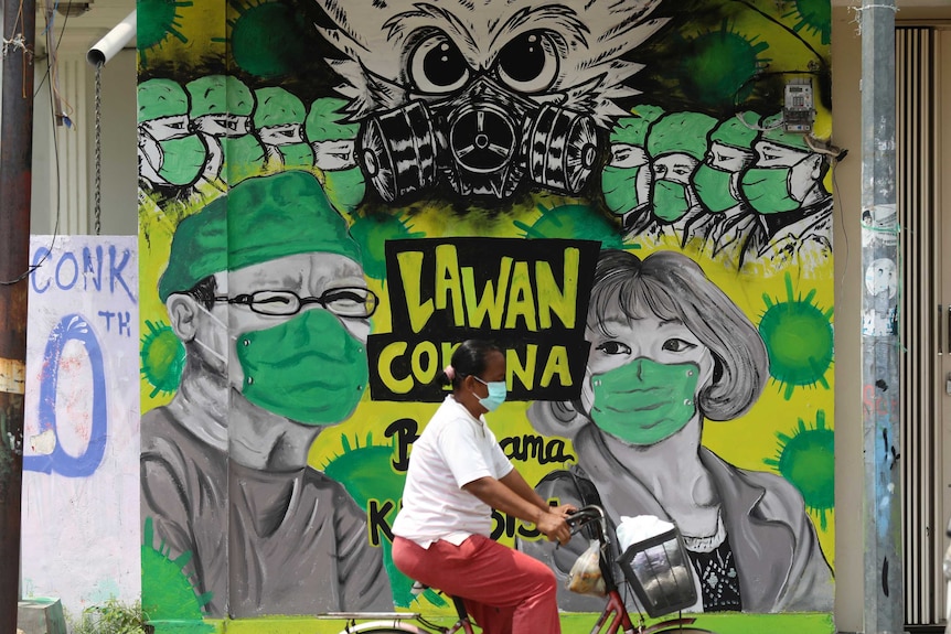 A woman rides her bicycle past a coronavirus awareness mural depicting medical workers in Indonesia.