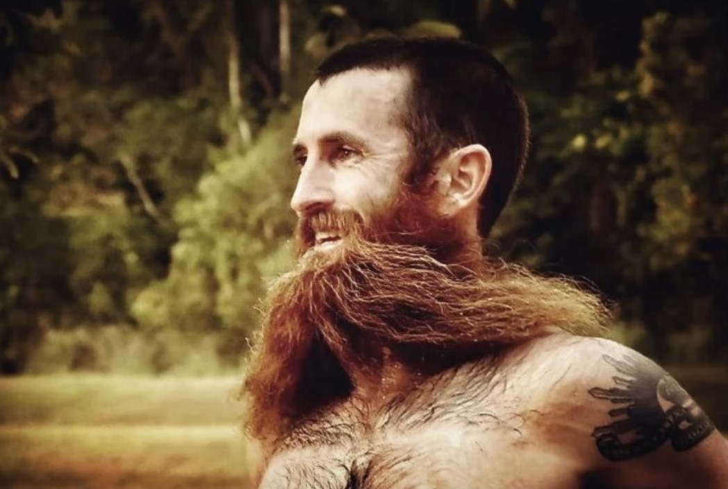 A man with short dark brown hair and brown, ginger beard flowing in the wind.