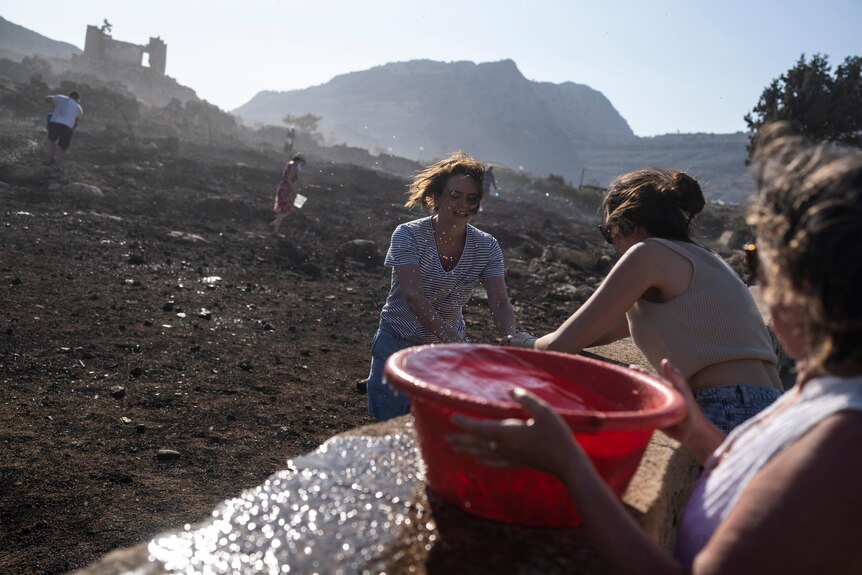 two women help an older greek lady full buckets of water 