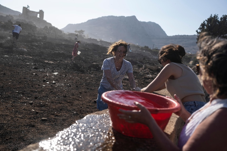 two women help an older greek lady full buckets of water 