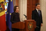 A middle-aged white woman with short brown hair speaks at a lectern while a middle-aged white man in a suit stands beside her.