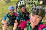 Three women in cycling gear sitting down with their mountain bikes behind them