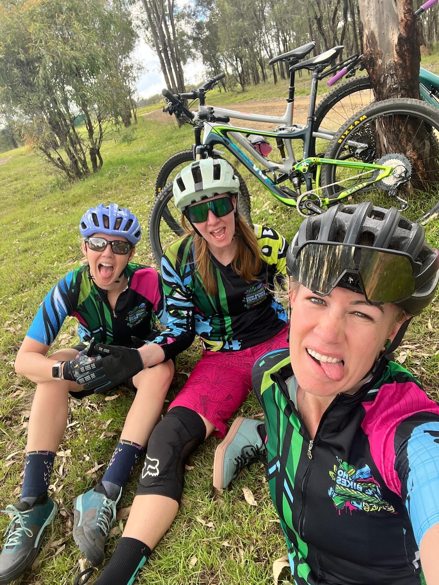 Three women in cycling gear sitting down with their mountain bikes behind them