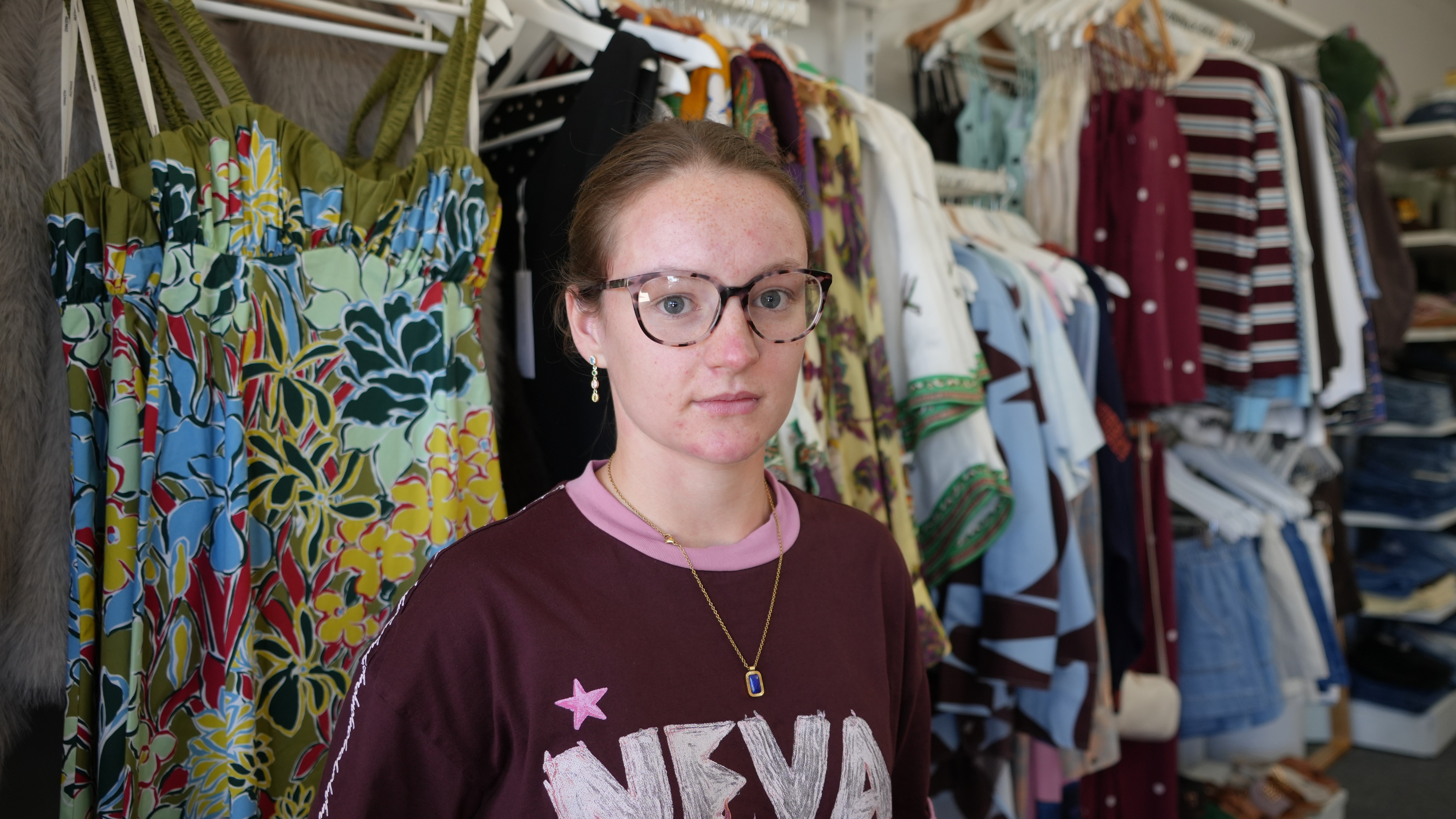 Orange haired young woman with a maroon top and glasses looks seriously at camera, in front of racks of clothes in shop