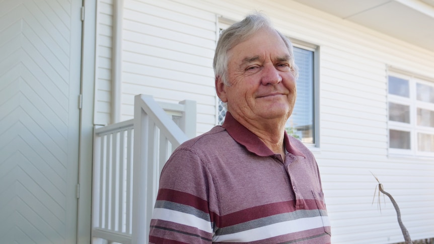 A man with short grey hair wearing a polo standing outside the front of a white weatherboard home