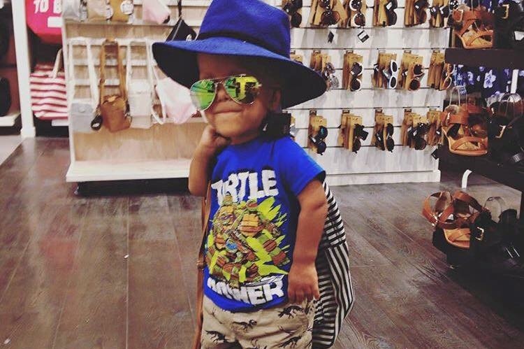 A young boy stands posing in a shop with bright oversized glasses and a hat on. He is smiling with a backpack over his shoulder.