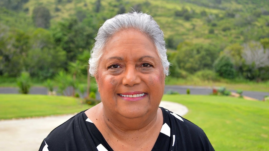 An Indigenous woman with white hair smiles in front of a grassy hill