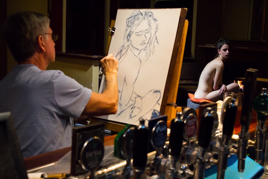 Man behind easel drawing a nude woman figure in pub venue.