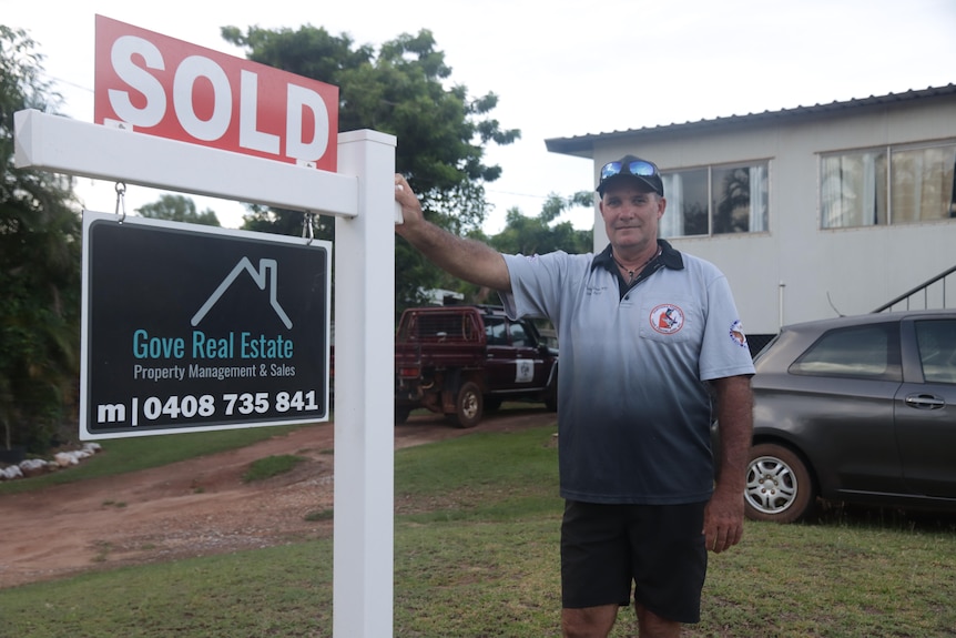A photo of Shaun Canobie standing next to sold house sign with two cars in the background and the house