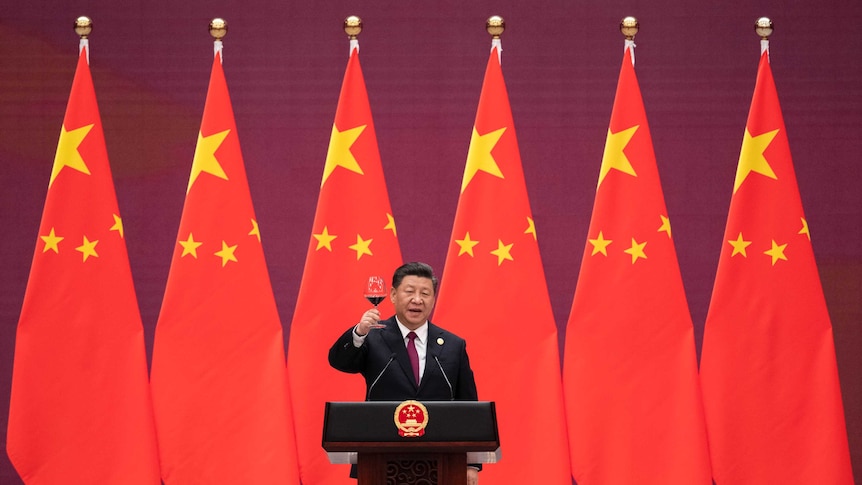 Xi Jinping stands in front of nine large Chinese flags as he toasts a glass of red wine standing behind an official lectern.