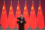 Xi Jinping stands in front of nine large Chinese flags as he toasts a glass of red wine standing behind an official lectern.