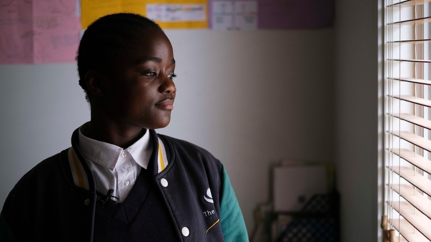 A teenage girl wearing school uniform looks out a window of her home