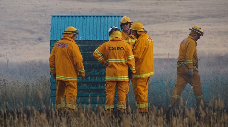 Five people from the CFA and CSIRO dressed in firefighting suits stand in a paddock besides a small blue shed.