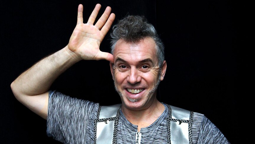 The performer, and man with salt and pepper hair and wearing a silver costume, smiles and waves into the camera.