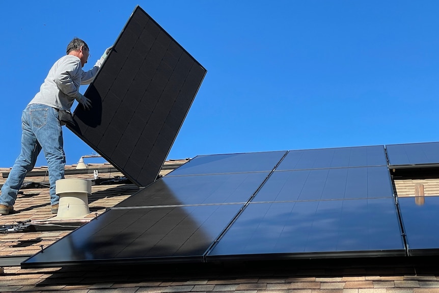 A worker places a solar panel on a roof next to those already in place.
