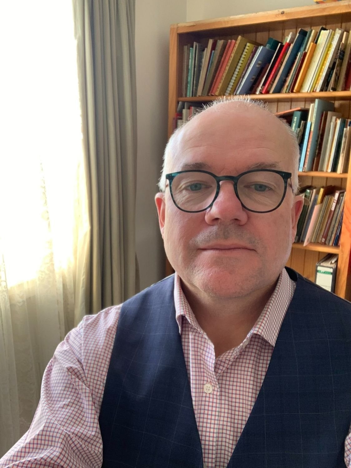 A bald Caucasian man with glasses takes a selfie in front of a bookshelf