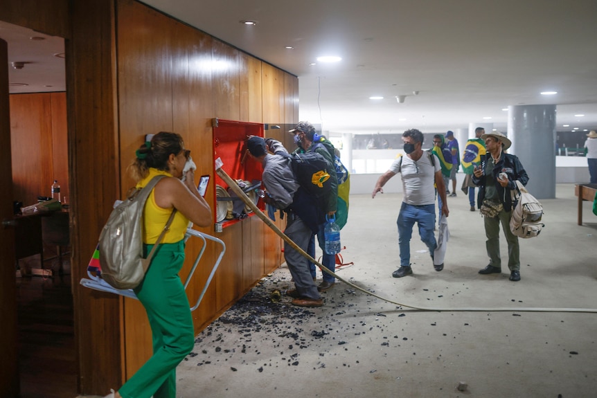 A woman walks out of an office as men smash a glass cabinet. 