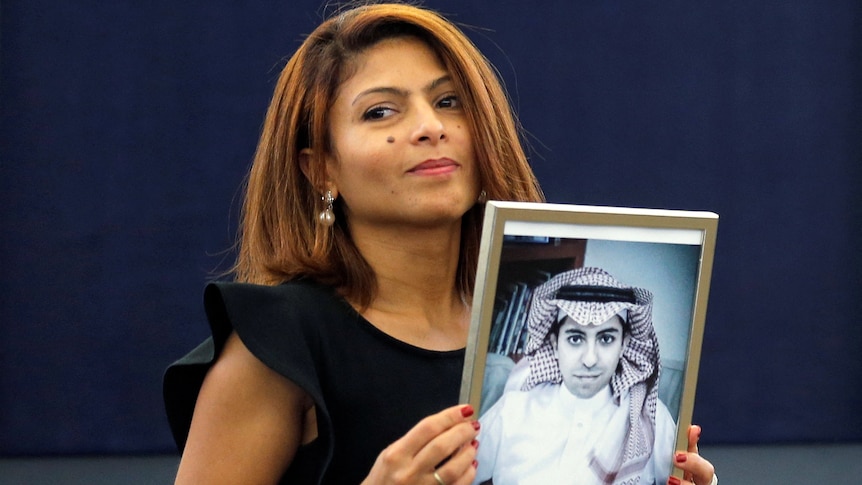 a woman in a black dress looks to the side as she holds up framed photo of her husband in white clothes and a keffiyeh 