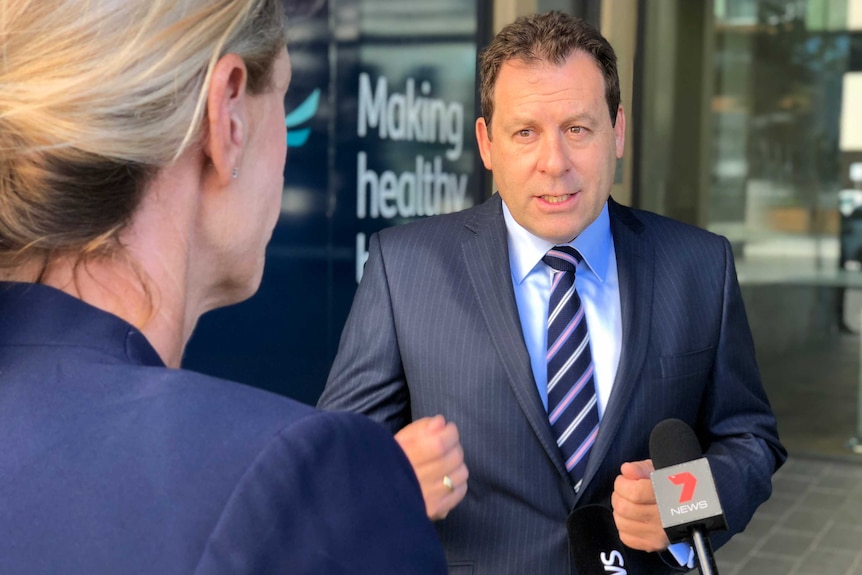 HBF CEO John Van Der Wielen in a blue suit seen over the shoulder of a blonde-haired female reporter.