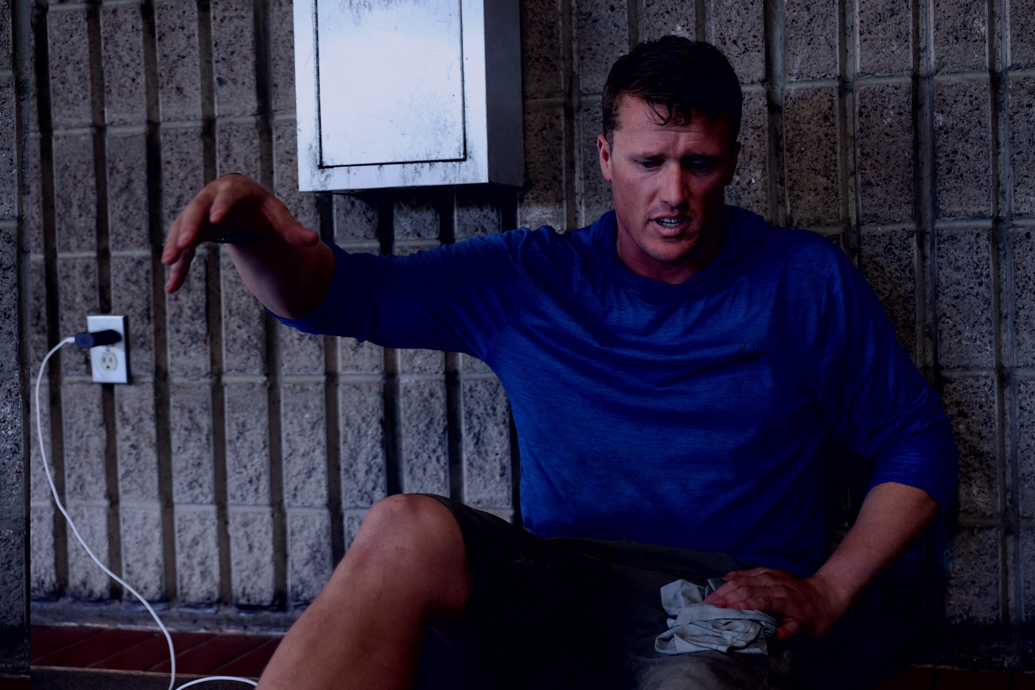 A young man in an athletic shirt sits on the ground and gestures as he speaks. His phone is plugged into the wall beside him.