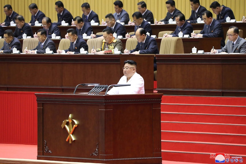Kim Jong Un delivers an opening speech at a conference as dozens of men in suits are seated around him at desks.