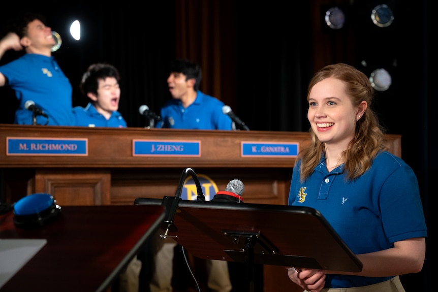 A film still of Alex Frias, Ben Wang, Mahi Alam and Angourie Rice celebrating a debating tournament, Rice behind a lectern.