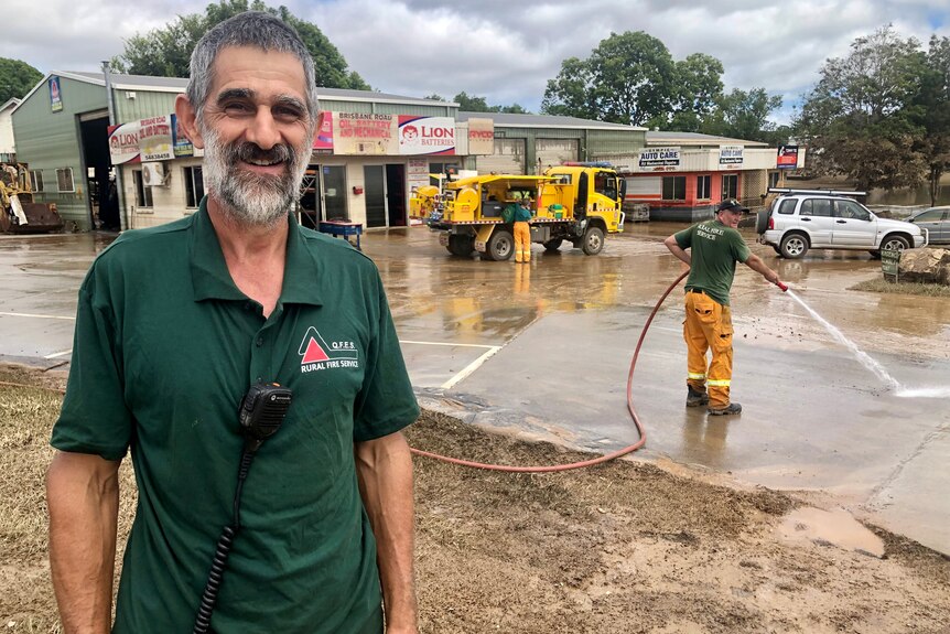 a man stands smiling at the camera while someone hoses cement in the background