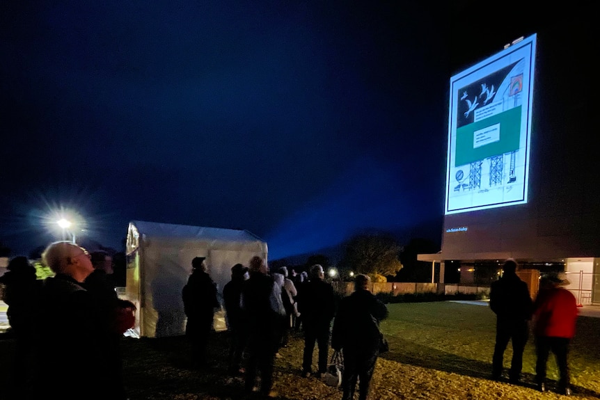 A crowd gathers at night to see a projection of art work on the side of a building
