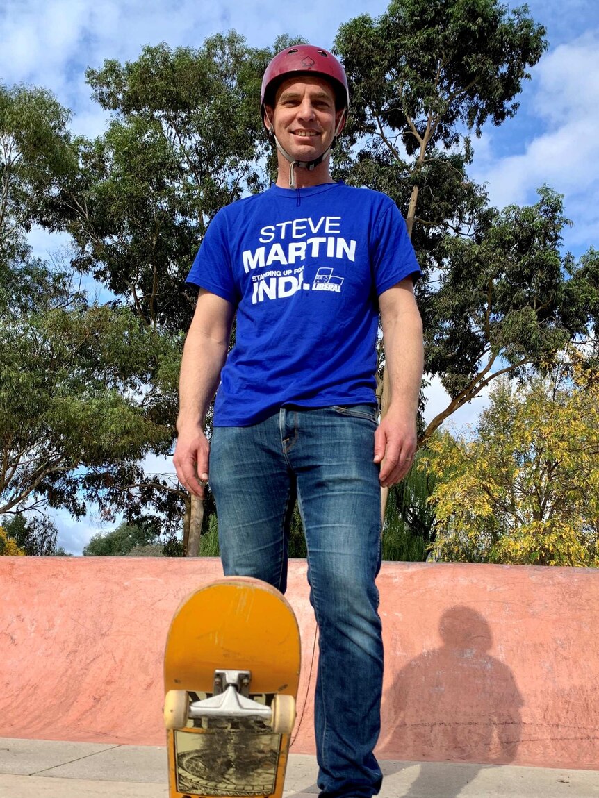 A man wearing a blue Liberal-branded shirt saying Steve Martin stands at the top of a skate park, with skateboard.