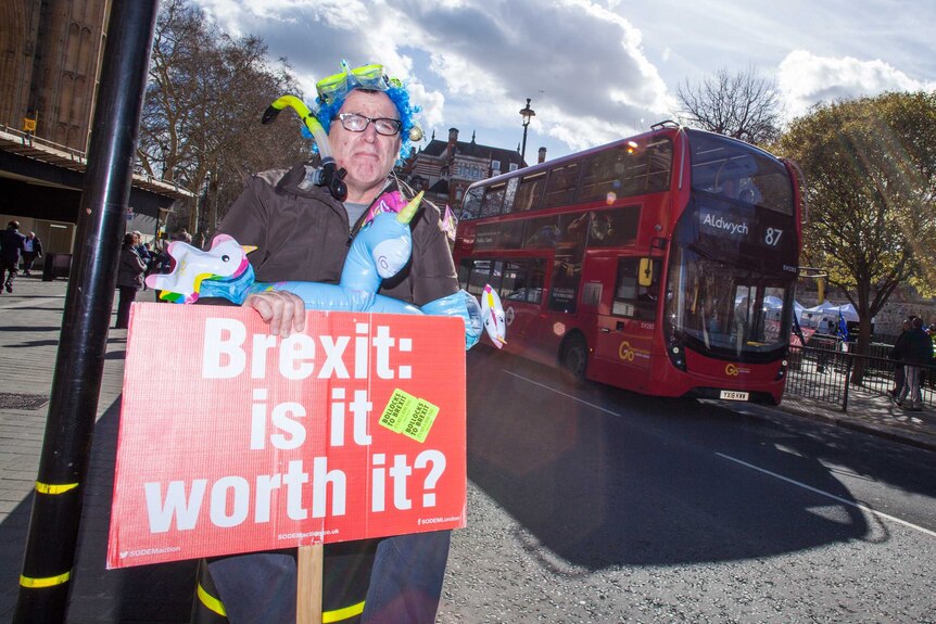 A man wearing a snorkel and pool toy holds a Brexit protest sign while standing in front of a passing double-decker bus.