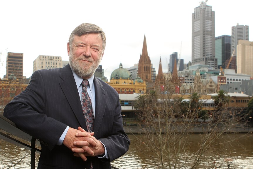 A man wearing a grey suit stands by the Yarra River. The Melbourne skyline is visible in the background.