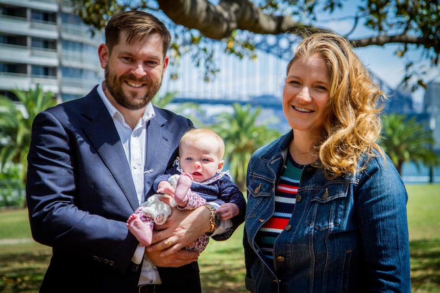 A man and a woman with a baby smiling at the camera in a park