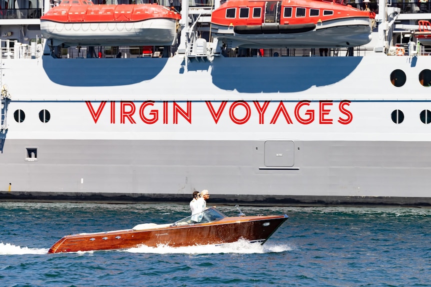 Two people on a brown speedboat next to a cruise ship reading Virgin Voyages.