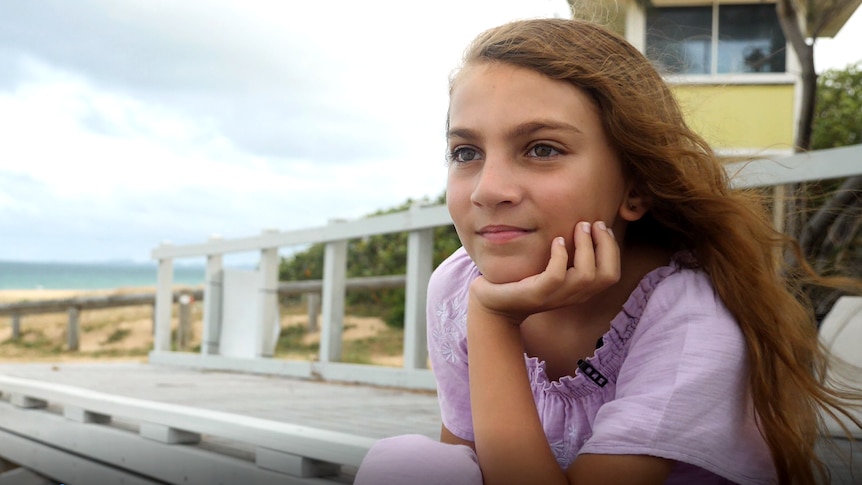 a young girl with long hair sits on a park bench, looking off into the distance with the beach behind her