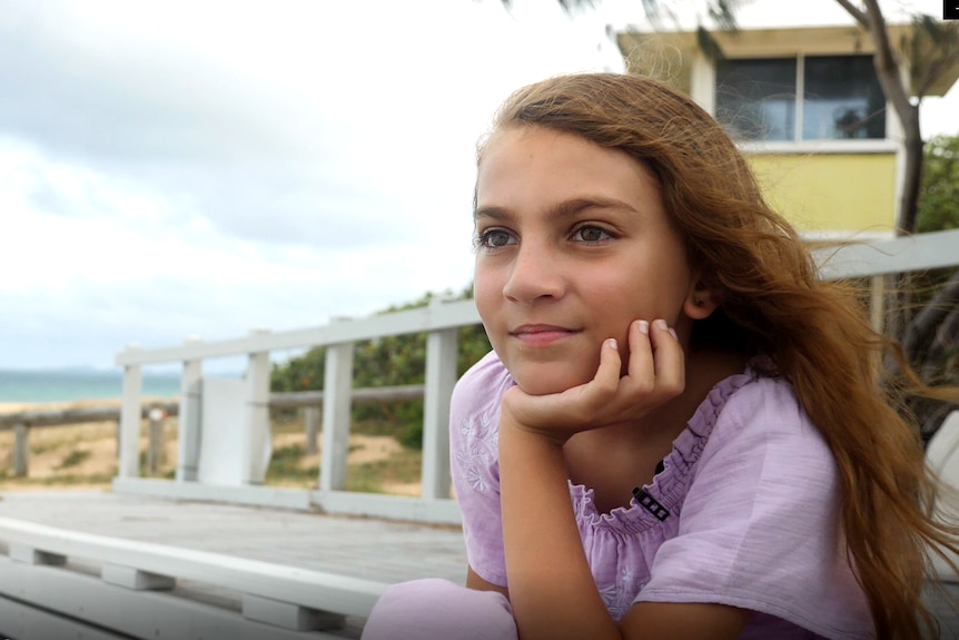 a young girl with long hair sits on a park bench, looking off into the distance with the beach behind her