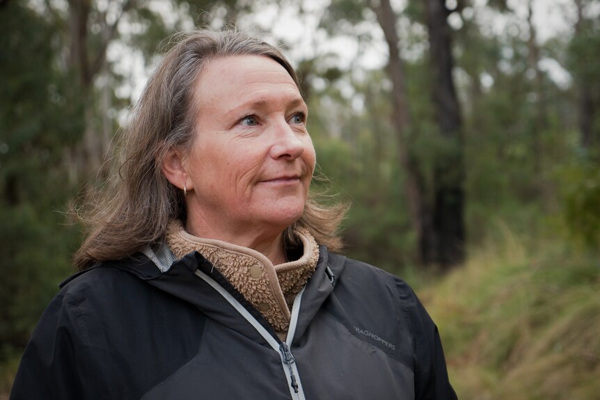 A woman with grey hair looks to the right of the camera with a slight smile while standing in bushland.