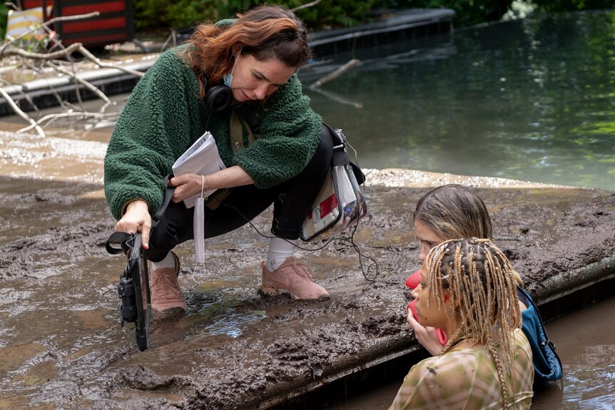 Woman with movie equipment talks to two women submerged up to their chests in dark muddy water.
