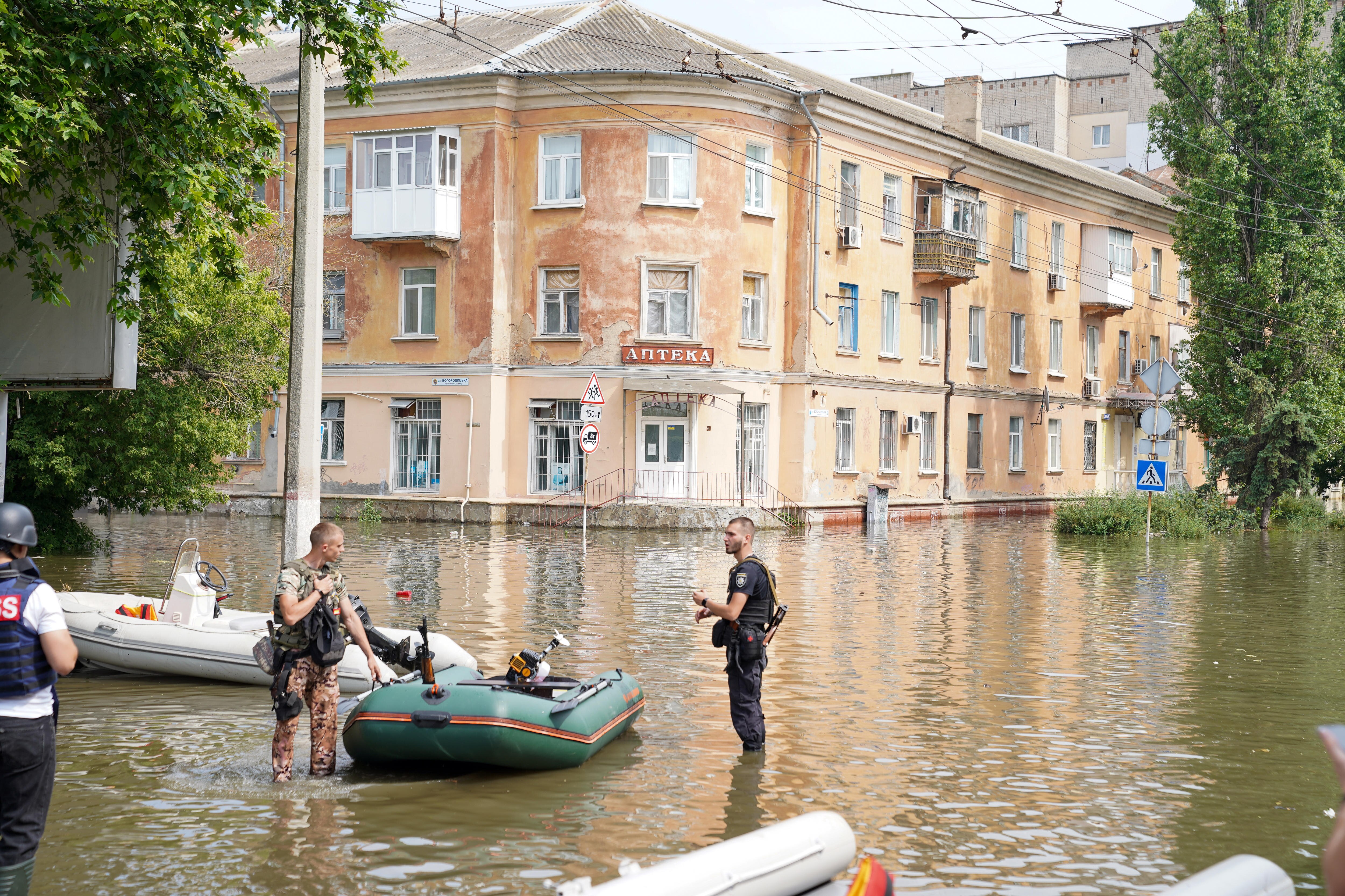Two men in uniform stand knee-deep in floodwater, talking as they handle a small green dingy