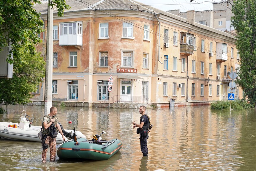 Two men in uniform stand knee-deep in floodwater, talking as they handle a small green dingy