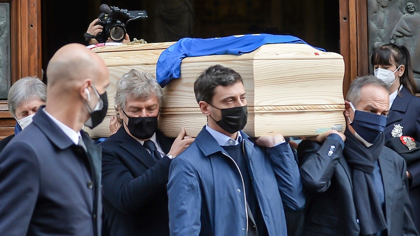 A group of men wearing dark blue coats carry a wooden coffin with a blue shirt on top