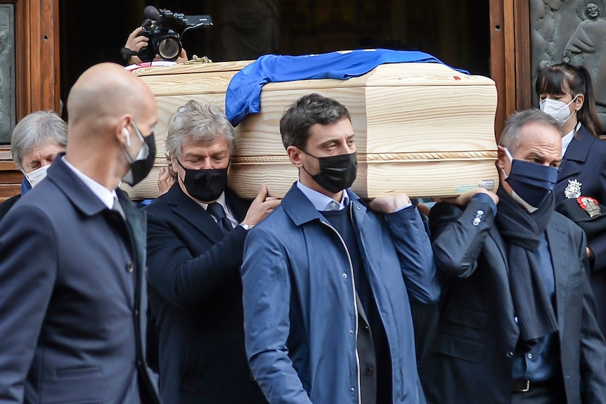 A group of men wearing dark blue coats carry a wooden coffin with a blue shirt on top